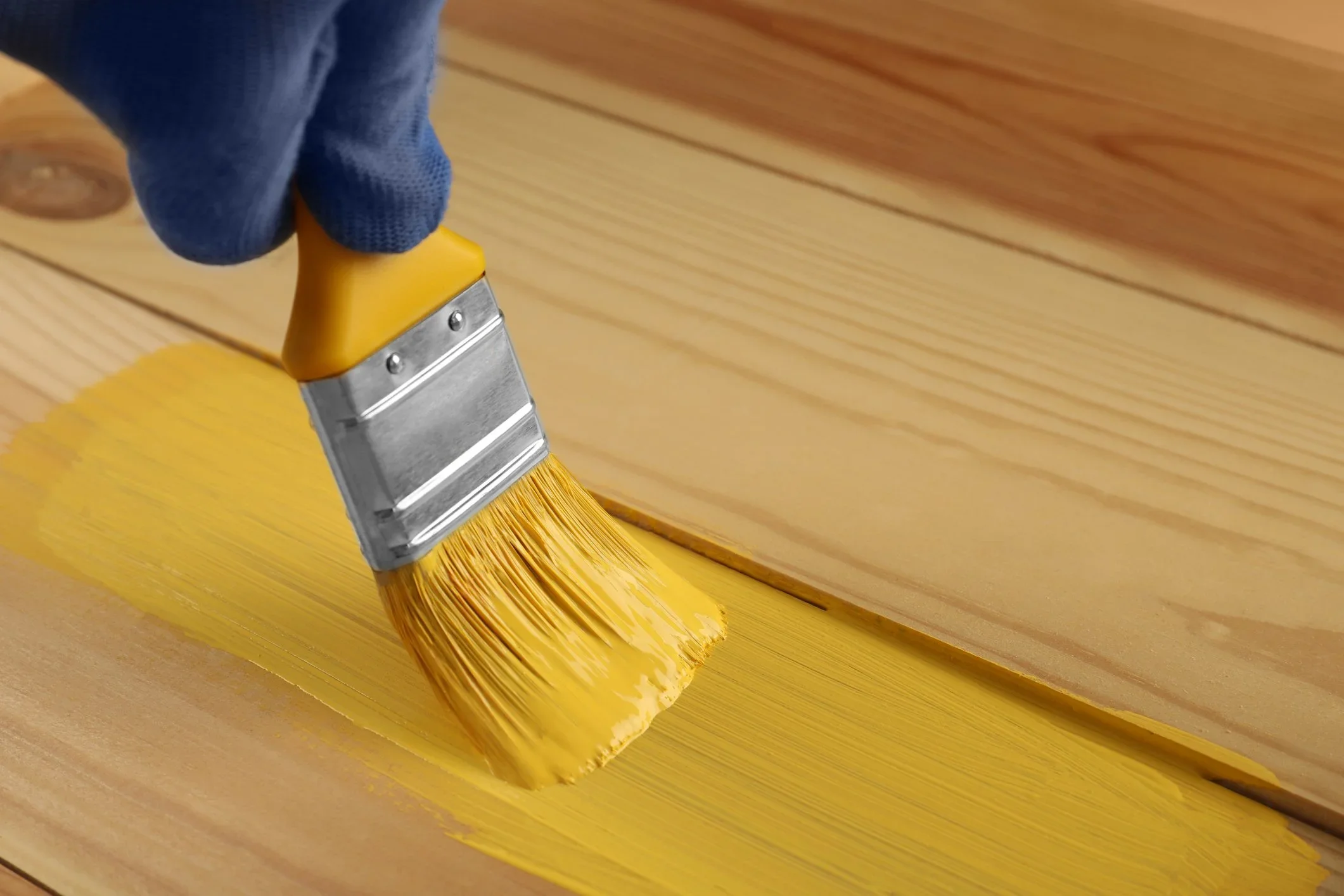 Gloved hand applying water based yellow paint to timber boards with a wide brush