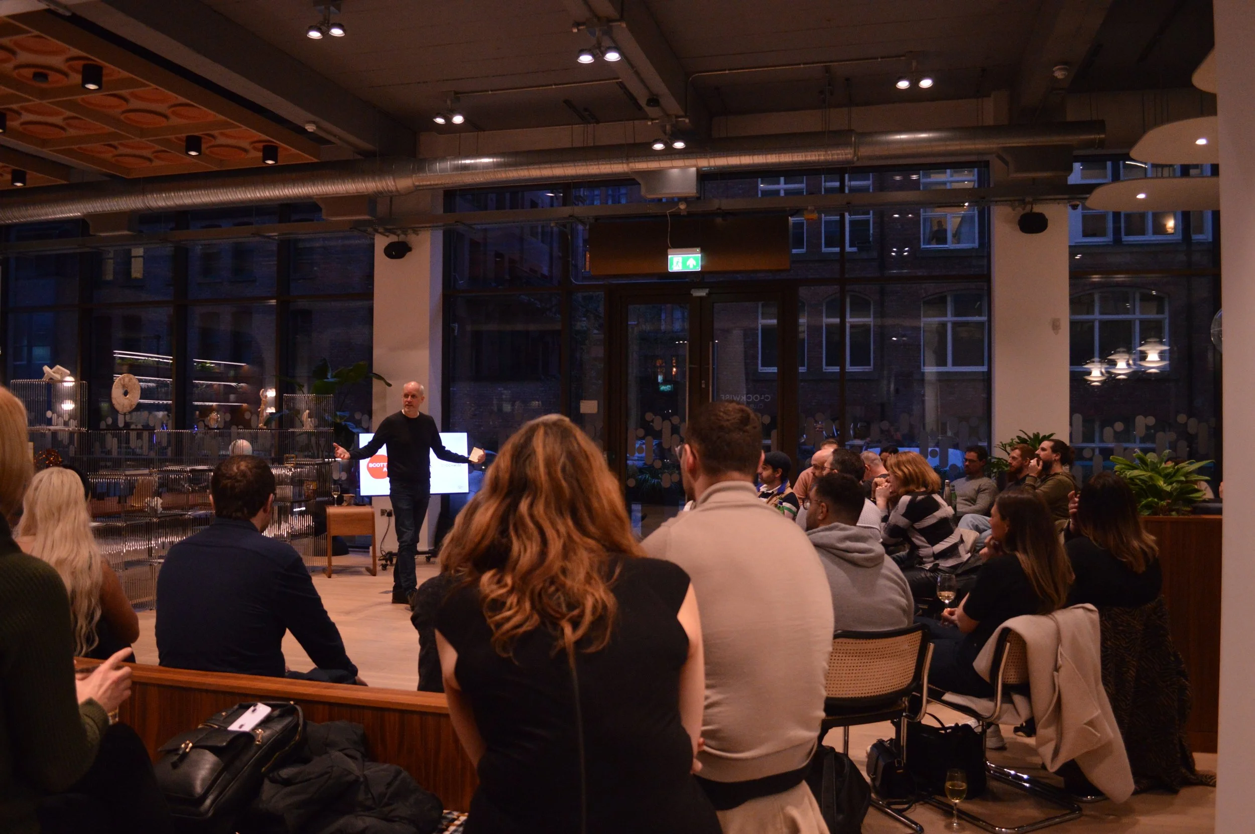 Martin Dangerfield, hosting a trumanchester event, to an audience in a modern, dimly lit conference room with large windows. The audience is seated and listening attentively.