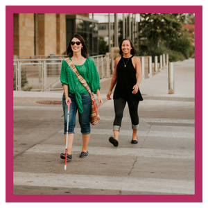 Two people walking across a street. 1 with cane.