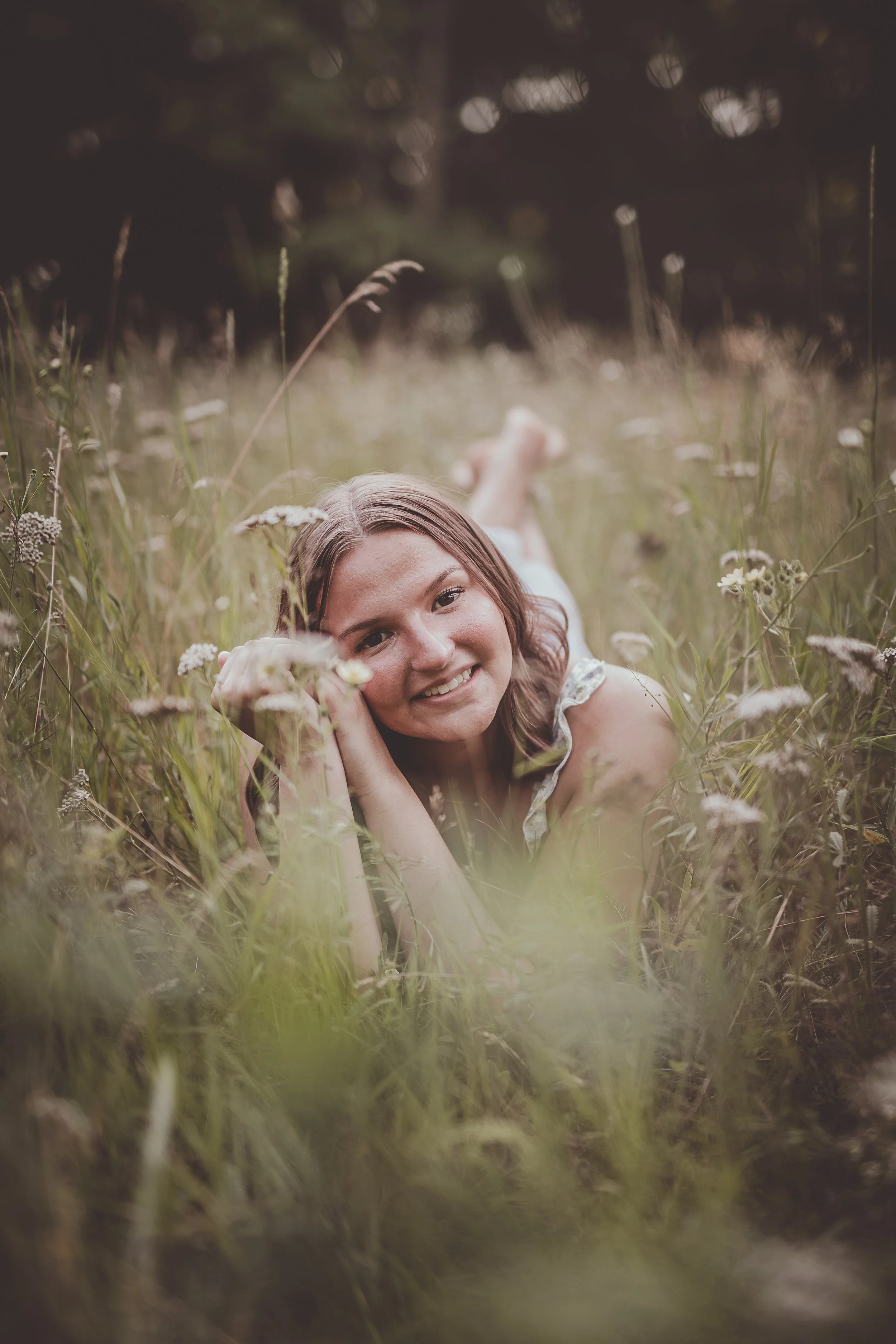 A woman lying in a grassy field surrounded by wildflowers, smiling and looking at the camera.