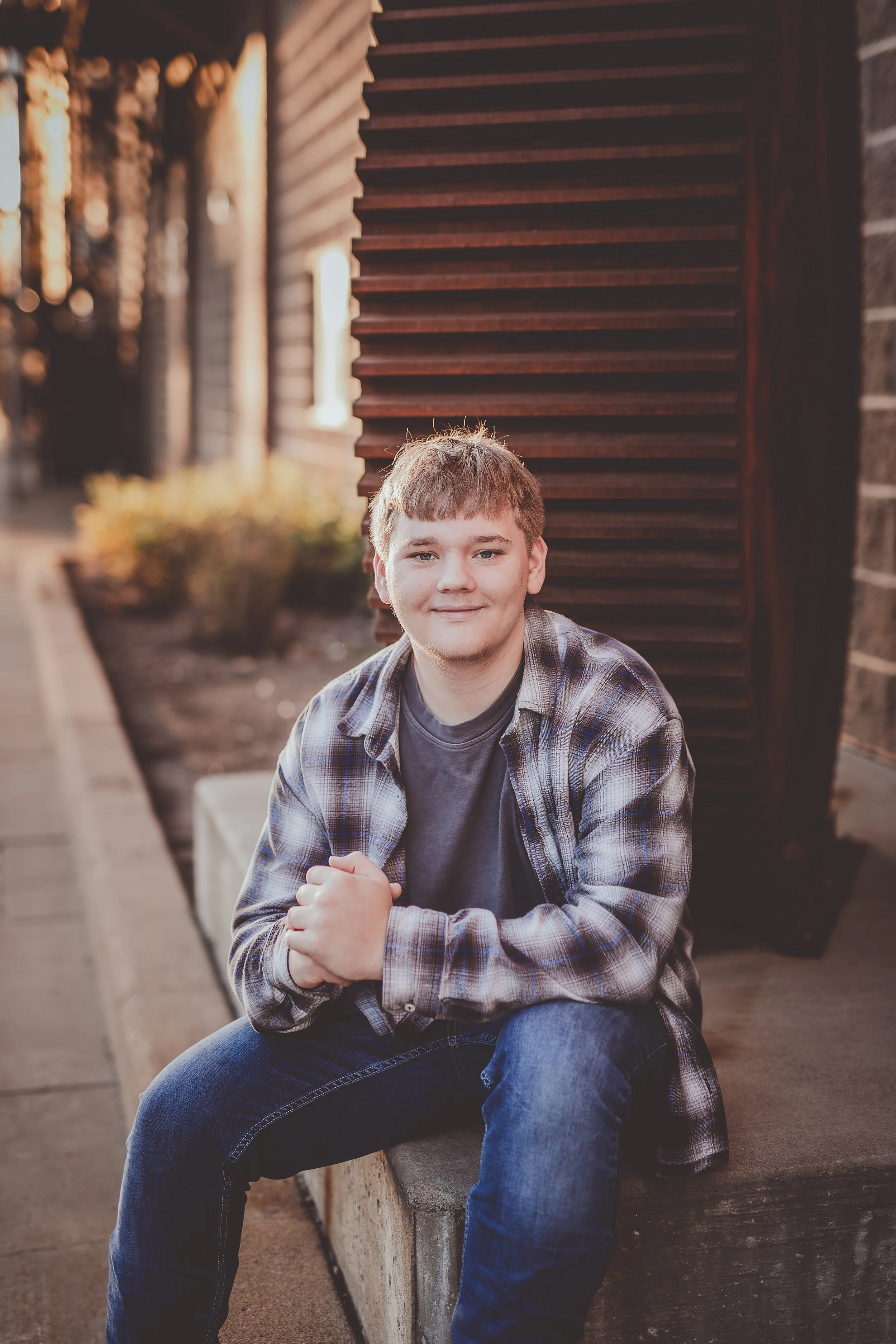 A young man with short light brown hair, wearing a plaid shirt over a gray t-shirt and blue jeans, sitting on a concrete bench outdoors, smiling, with a wooden wall and potted plants in the background.