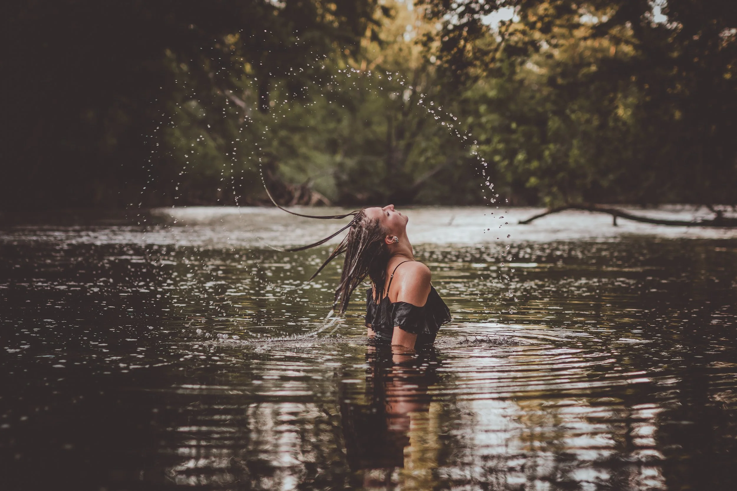 A woman with long wet hair is standing in a river, flipping her head back and splashing water into the air, surrounded by trees in a natural setting.