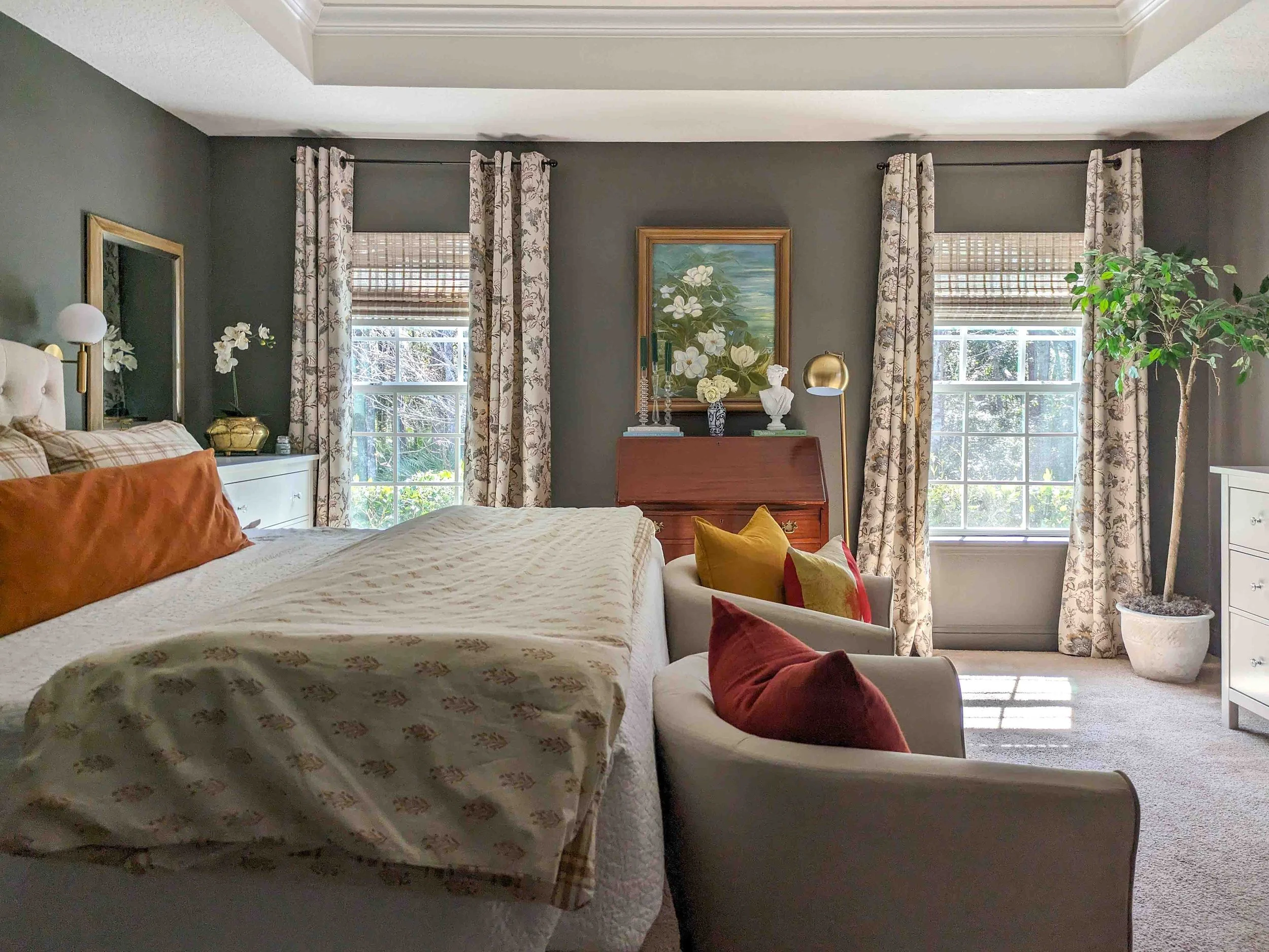 A cozy, traditional bedroom features dark charcoal walls, floral patterned curtains framing two windows, and a white bed accented with warm terracotta and mustard yellow pillows.