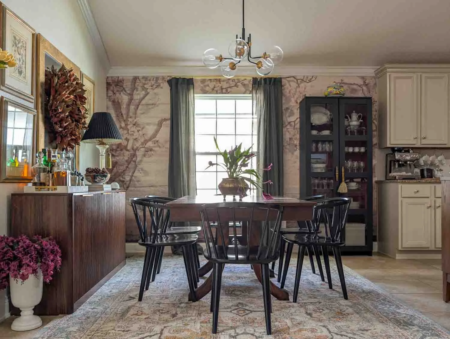 A formal dining room featuring a scenic mural wallpaper and dark green sheer curtains. A dark wood dining table is surrounded by black spindle chairs.