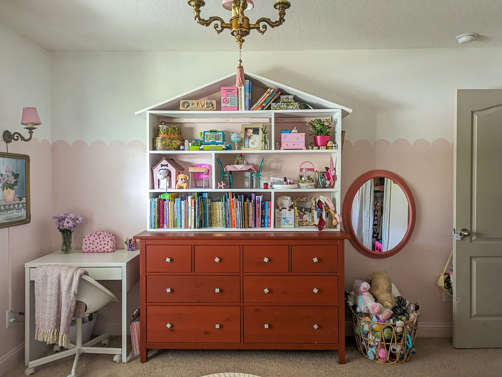 A whimsical child's room features a large terracotta dresser topped with a house-shaped bookshelf filled with toys and books, set against a wall with a soft pink scalloped paint detail and a matching round mirror.
