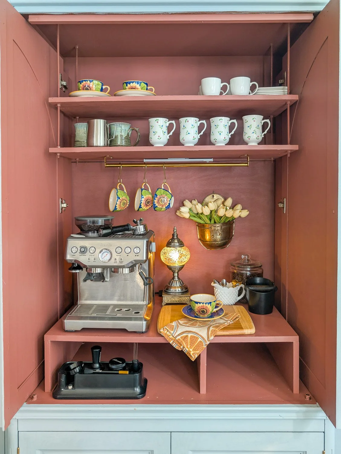 A dusty rose-painted hutch interior serves as a coffee bar, equipped with a stainless steel espresso machine, shelves of assorted mugs, a small glowing lamp, and a brass wall planter holding white tulips.