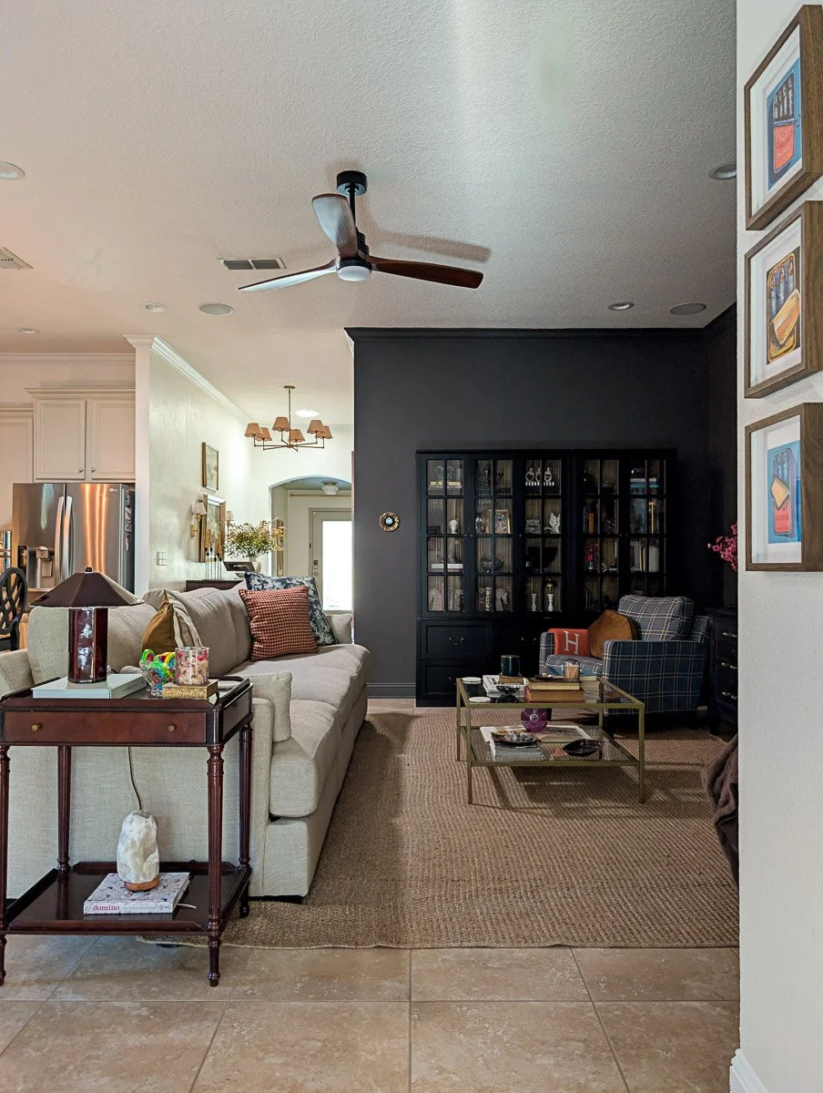 A cozy living room features a neutral sofa and plaid armchair arranged around a gold-framed coffee table, set against a dramatic dark green accent wall with a large black glass-door cabinet.