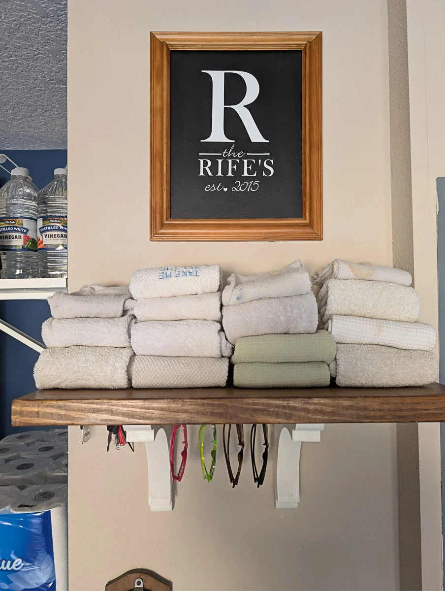 A wooden floating shelf holds stacks of white towels and features white brackets with sunglasses hanging underneath, positioned below a framed monogram sign.