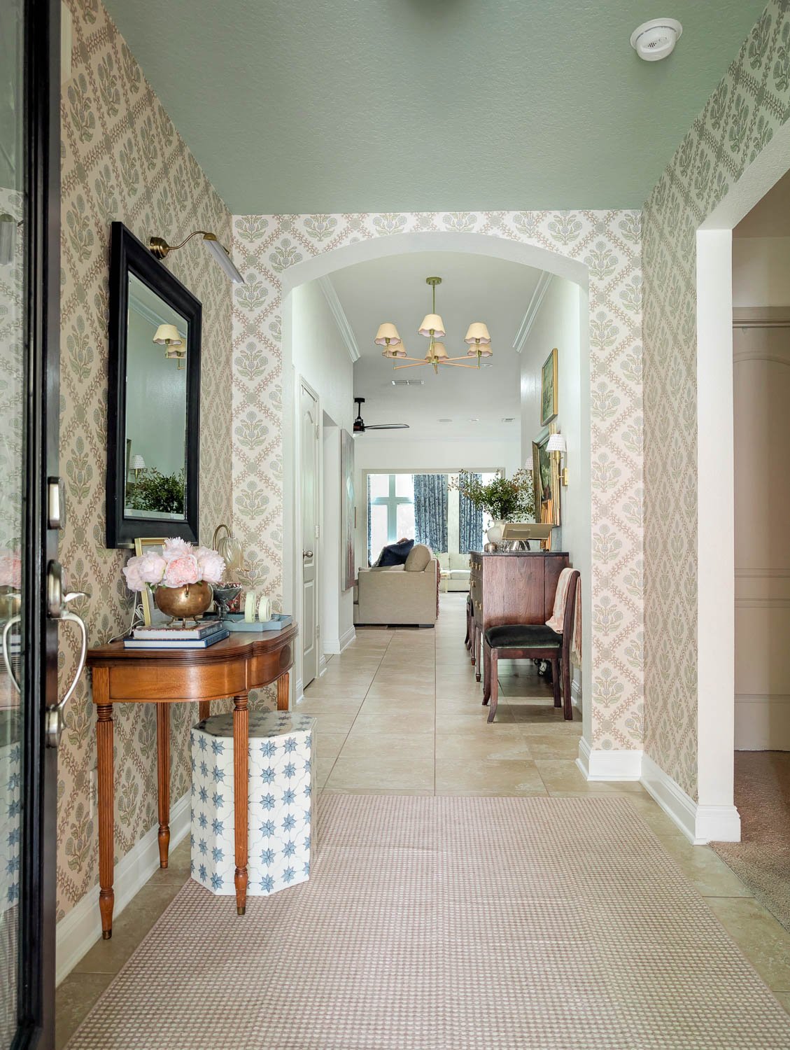 A moody traditional entryway features a large-scale floral wallpaper in muted tones, a vintage scalloped wood console table styled with heirloom art, and a floor-to-ceiling dark floral drape that complements the "Antique Jade" painted ceiling.
