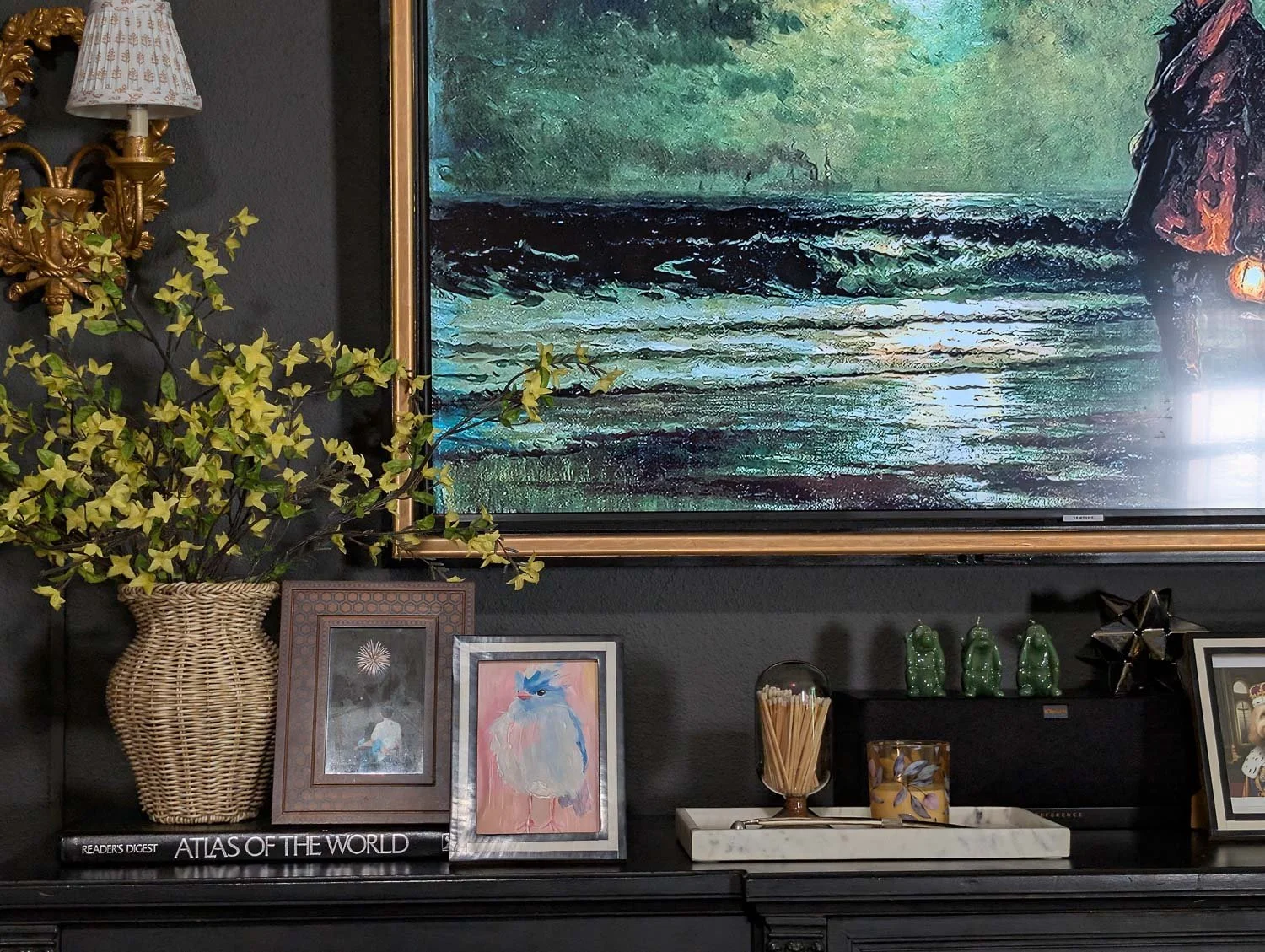 A warm wood media console styled for summer with a basket vase of vibrant yellow branches, leaning framed art, and a vintage-style radio against deep charcoal gray walls in a modern heritage living room.