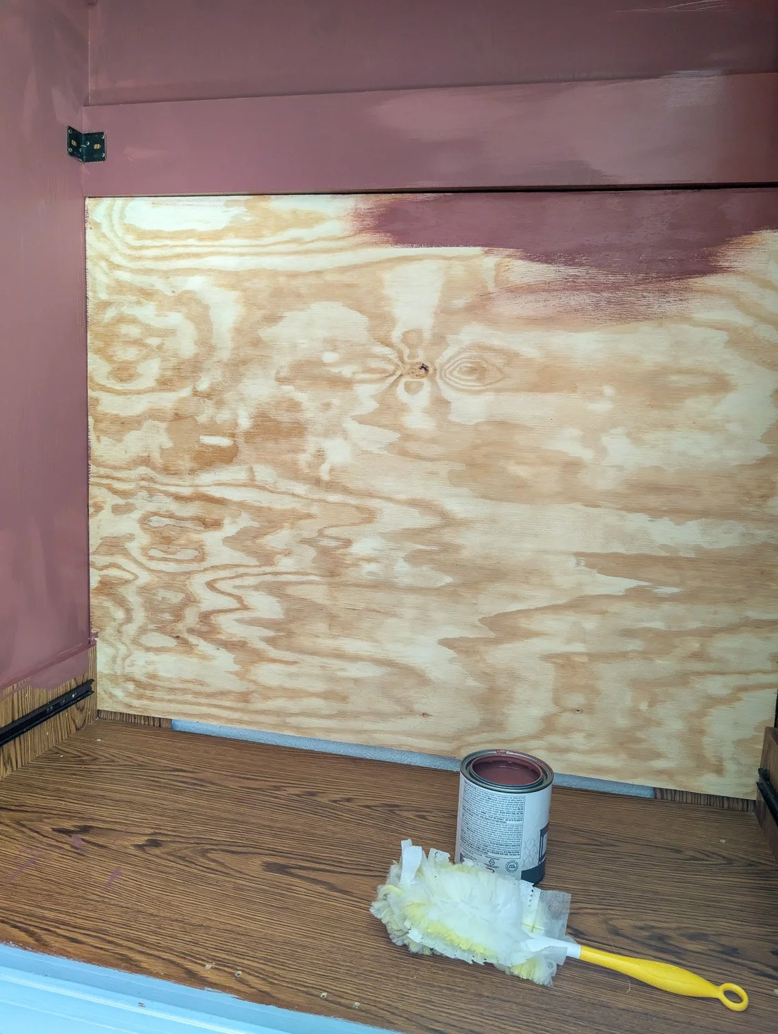 A close-up of a DIY plywood backing being installed and partially painted with dark red paint inside a wooden cabinet.