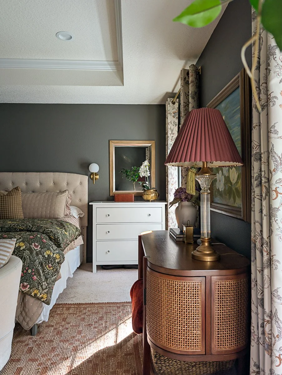 A cozy, traditional bedroom features a dark charcoal accent wall, a tufted beige headboard with floral bedding, and a vintage-style lamp with a pleated burgundy shade atop a cane-paneled wood console.