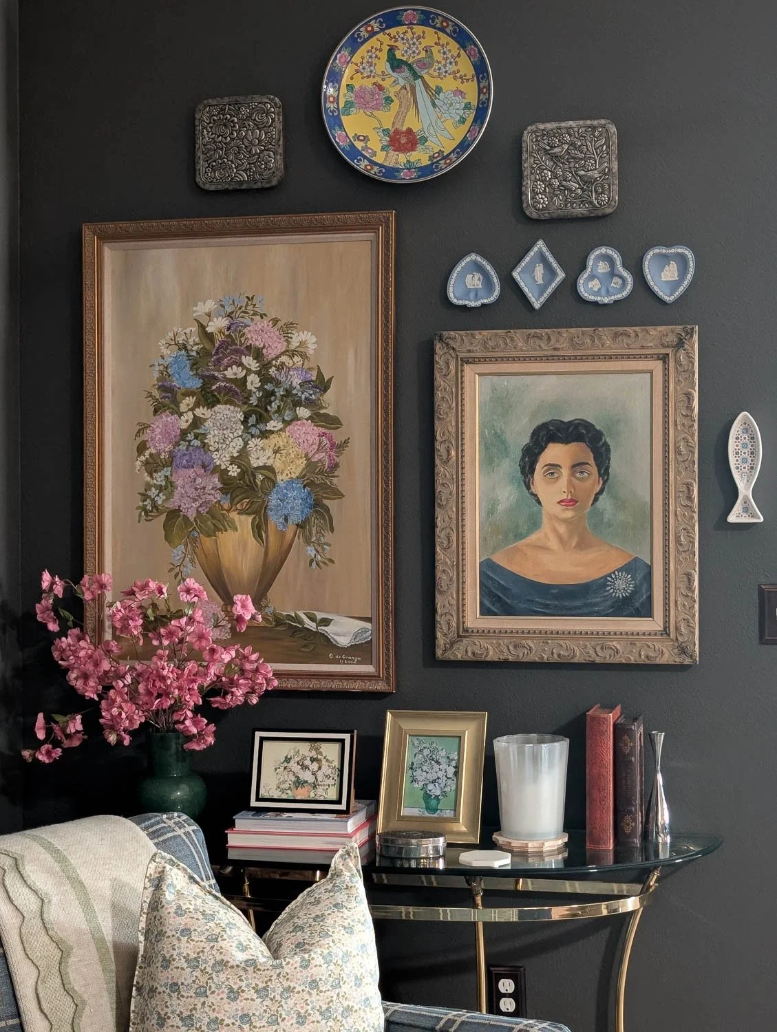A gallery wall styled against a dark charcoal-grey backdrop. The arrangement includes a large gold-framed floral oil painting, a smaller portrait of a woman, and various decorative plates and Wedgwood-style blue jasperware.