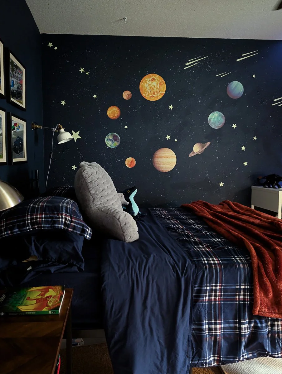 A cozy bedroom features a dark navy accent wall adorned with a colorful solar system mural and glowing stars, centered behind a bed with navy and plaid linens and a rust-colored throw blanket.