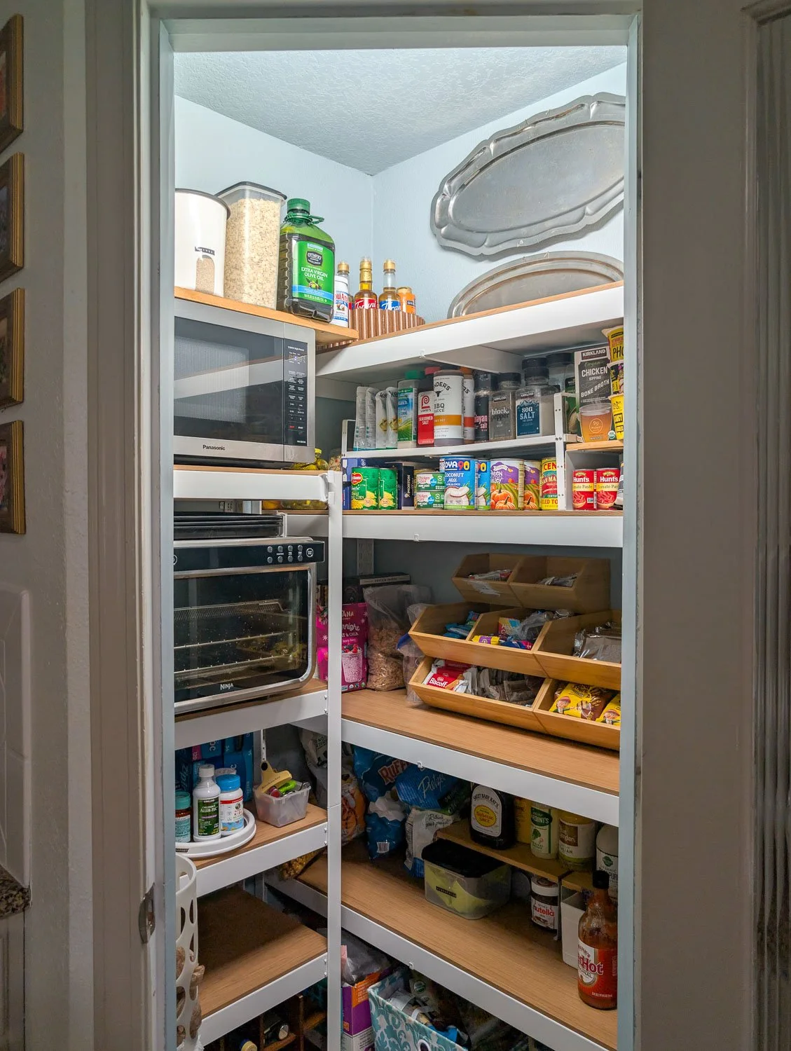 A beautifully organized pantry featuring white metal and wood shelving against light blue walls, accented by vintage silver pewter platters hung near the ceiling.