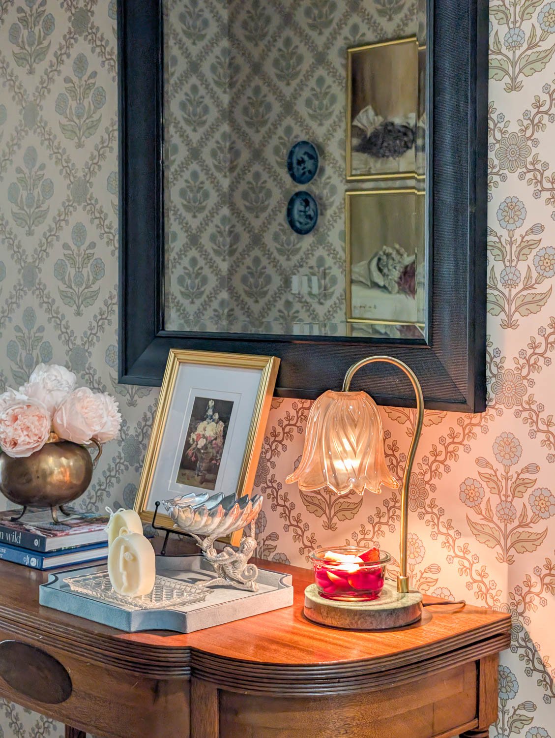 A floral-style glass candle warmer on a green marble base casting a warm glow over a simmer pot in a moody, wallpapered entryway.