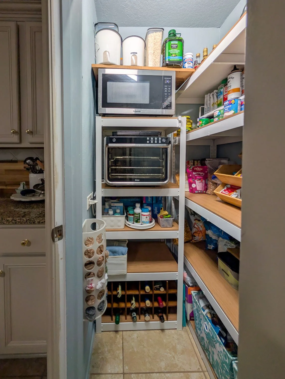 A narrow white metal shelving unit functioning as an appliance station inside a pantry, holding a microwave, a mini oven, and various household supplies.