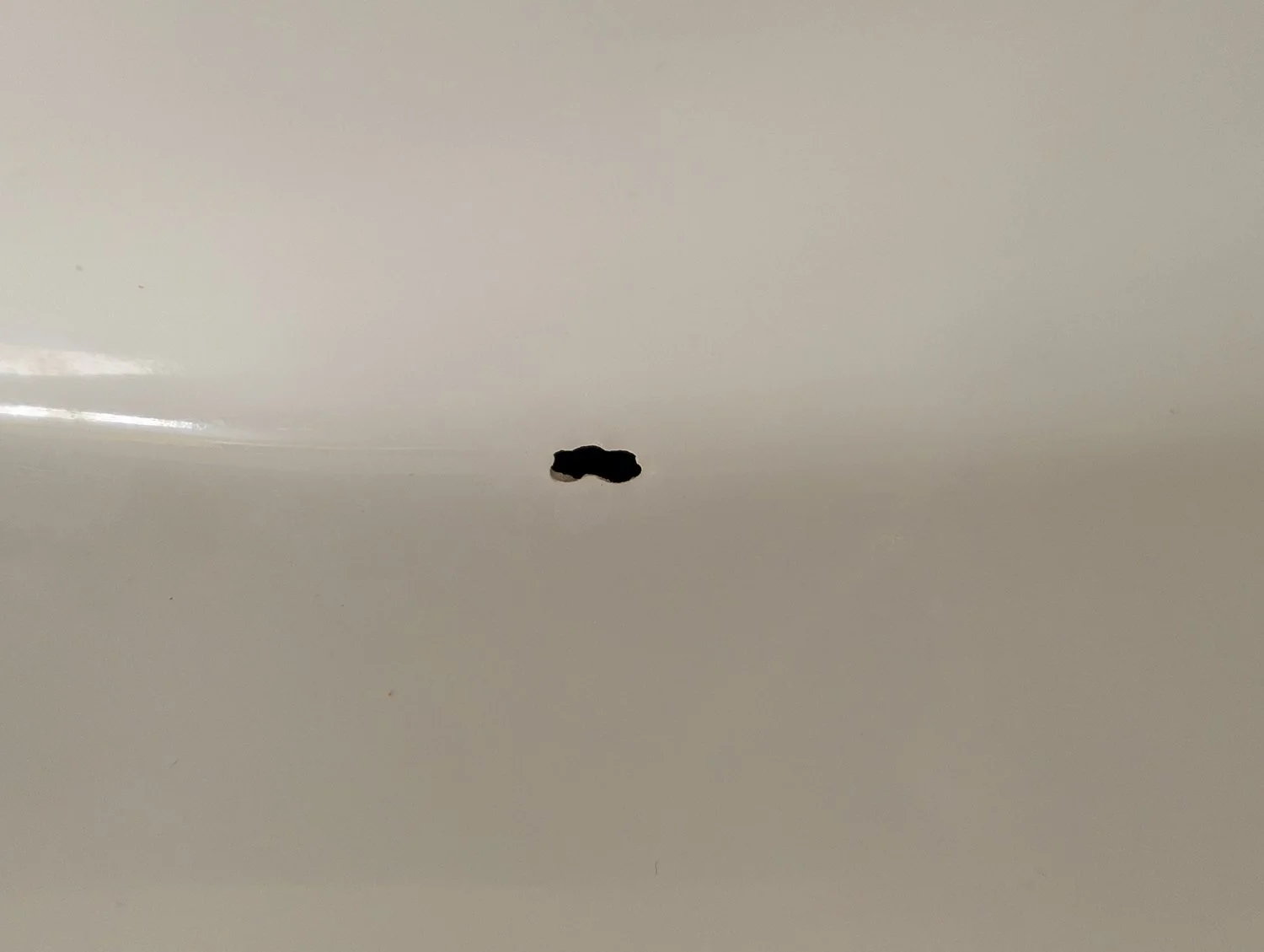 A close-up, top-down view of a small, dark, jagged hole in a smooth white porcelain bathroom sink.