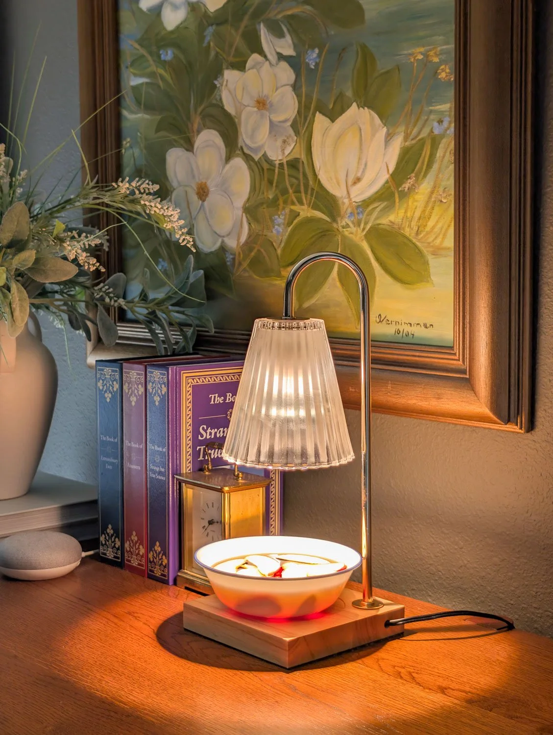 A ribbed glass candle warmer lamp positioned on a wooden desk next to a vase of greenery and a large magnolia painting.
