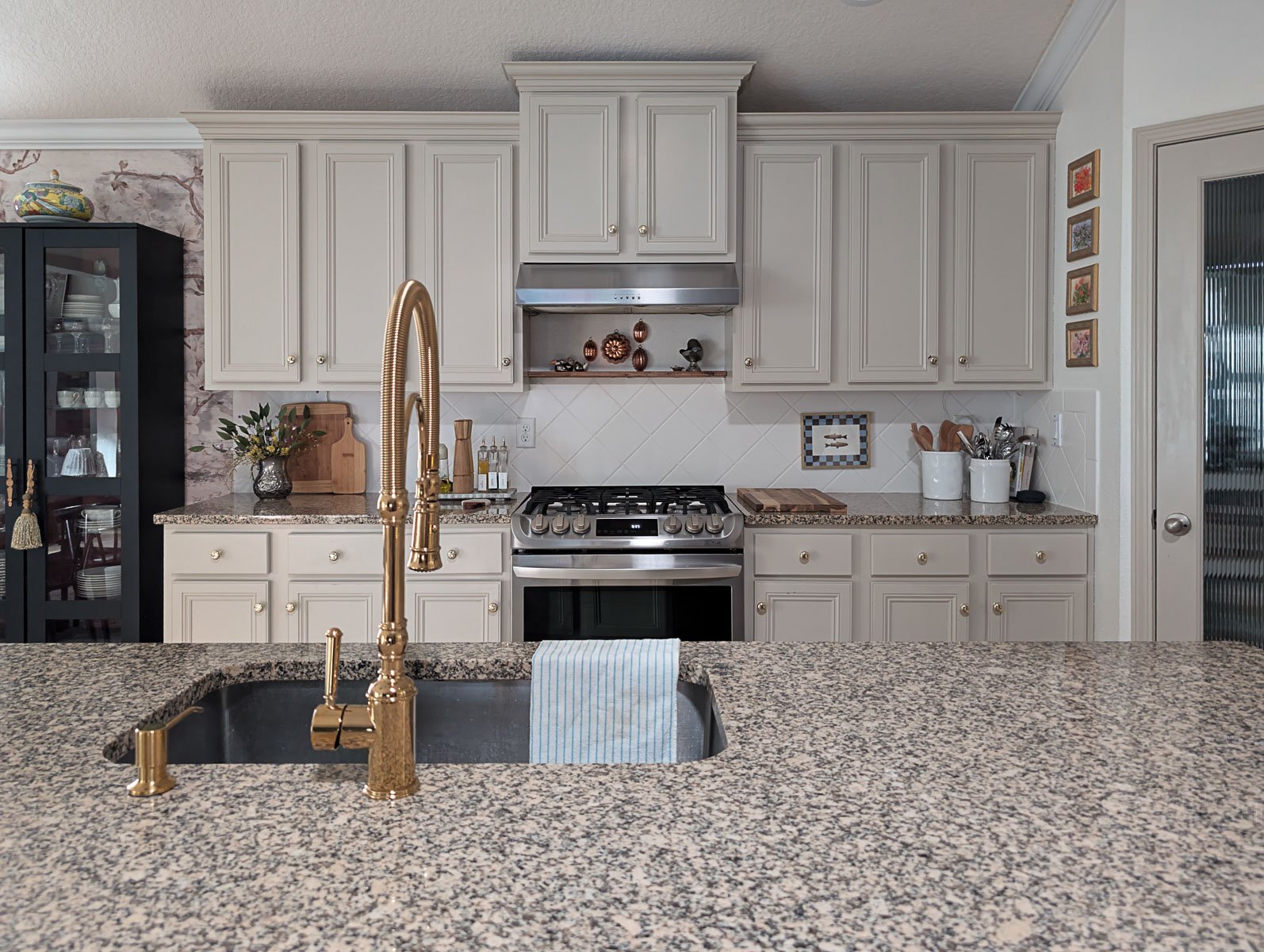 A modern kitchen features light beige cabinetry with gold hardware, a speckled granite island with a gold pull-down faucet, and a stainless steel gas range set against a white tiled backsplash.