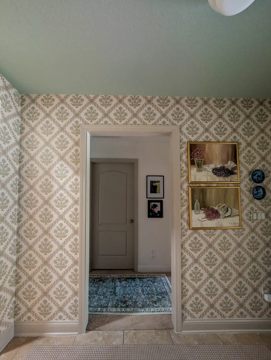A wide shot of a hallway featuring floral patterned wallpaper, a sage green ceiling, and a doorway framed with neutral-toned trim.