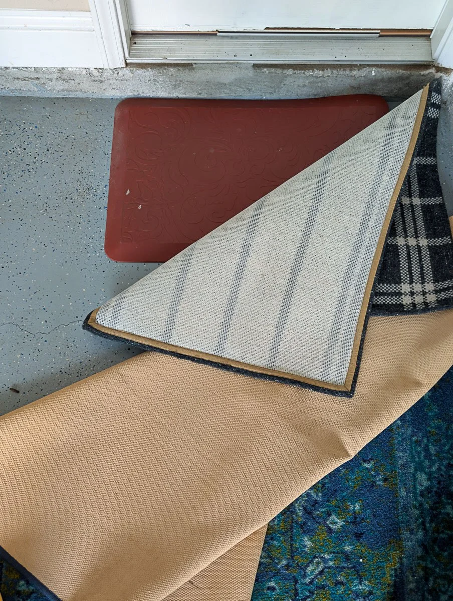 A pile of various floor mats and a blue patterned rug are shown being layered or sorted on a gray speckled garage floor.