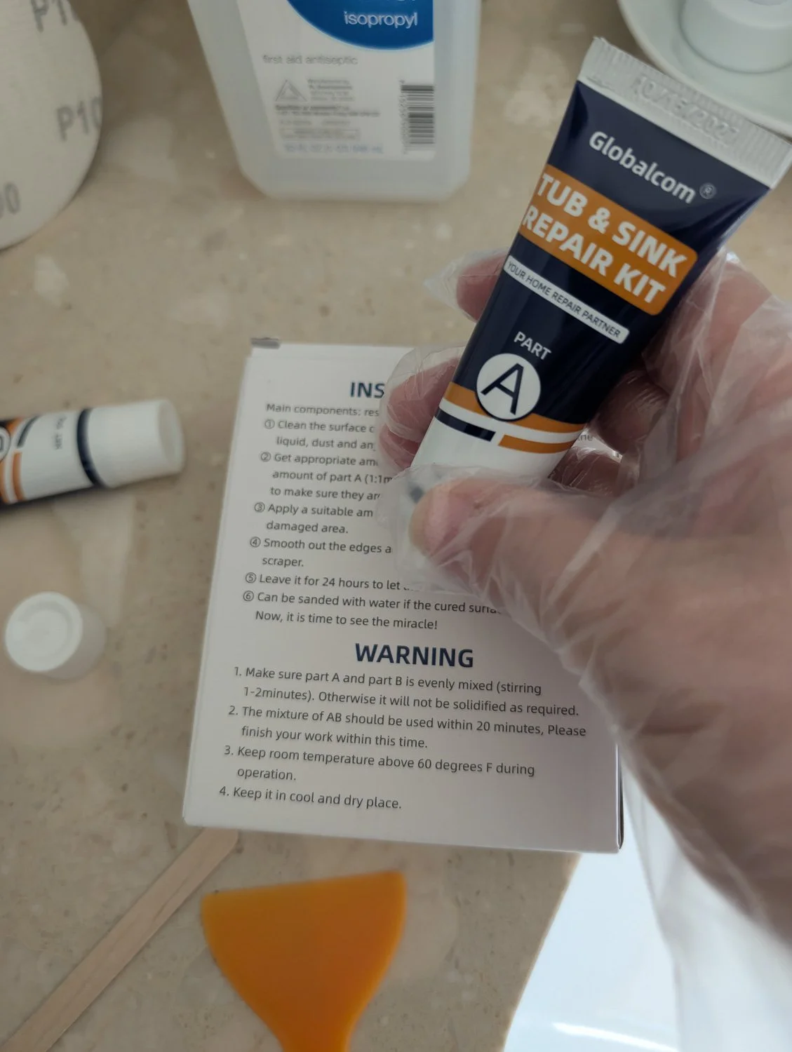 A hand wearing a clear protective glove holds a tube of "Part A" epoxy resin from a tub and sink repair kit, with instructions and a small orange spreader in the background.