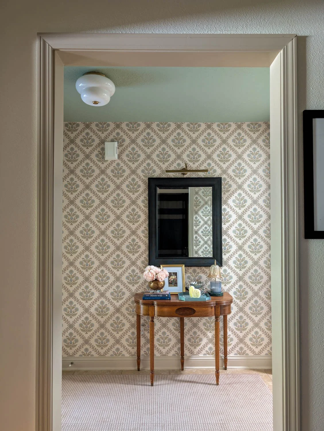 A view through a trimmed doorway showcasing a wooden entryway table decorated with a mirror, floral arrangements, and books against patterned wallpaper.