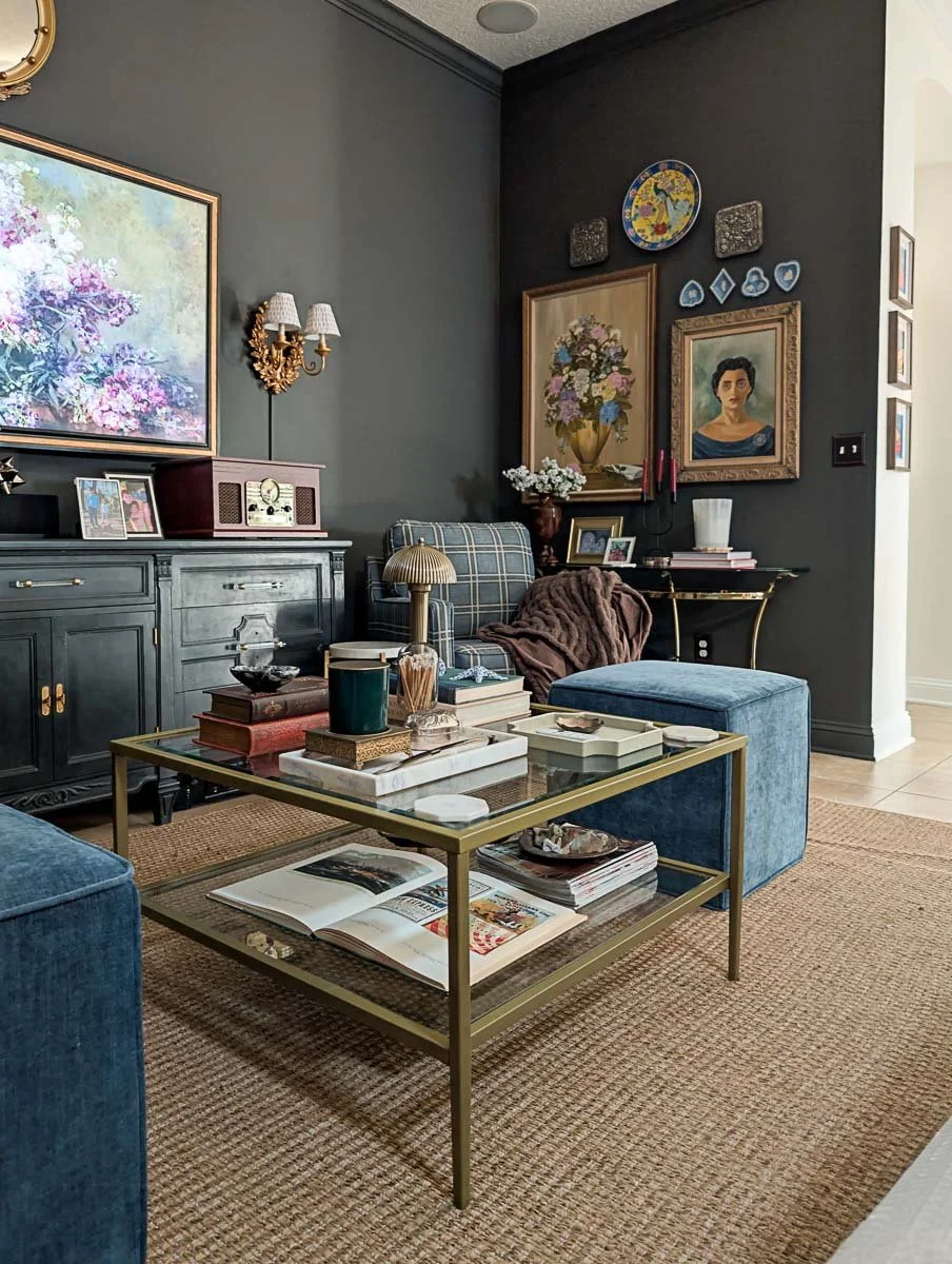 A moody, eclectic living room features dark green walls decorated with gold-framed artwork, blue plaid armchairs, and a gold-framed glass coffee table on a natural jute rug.