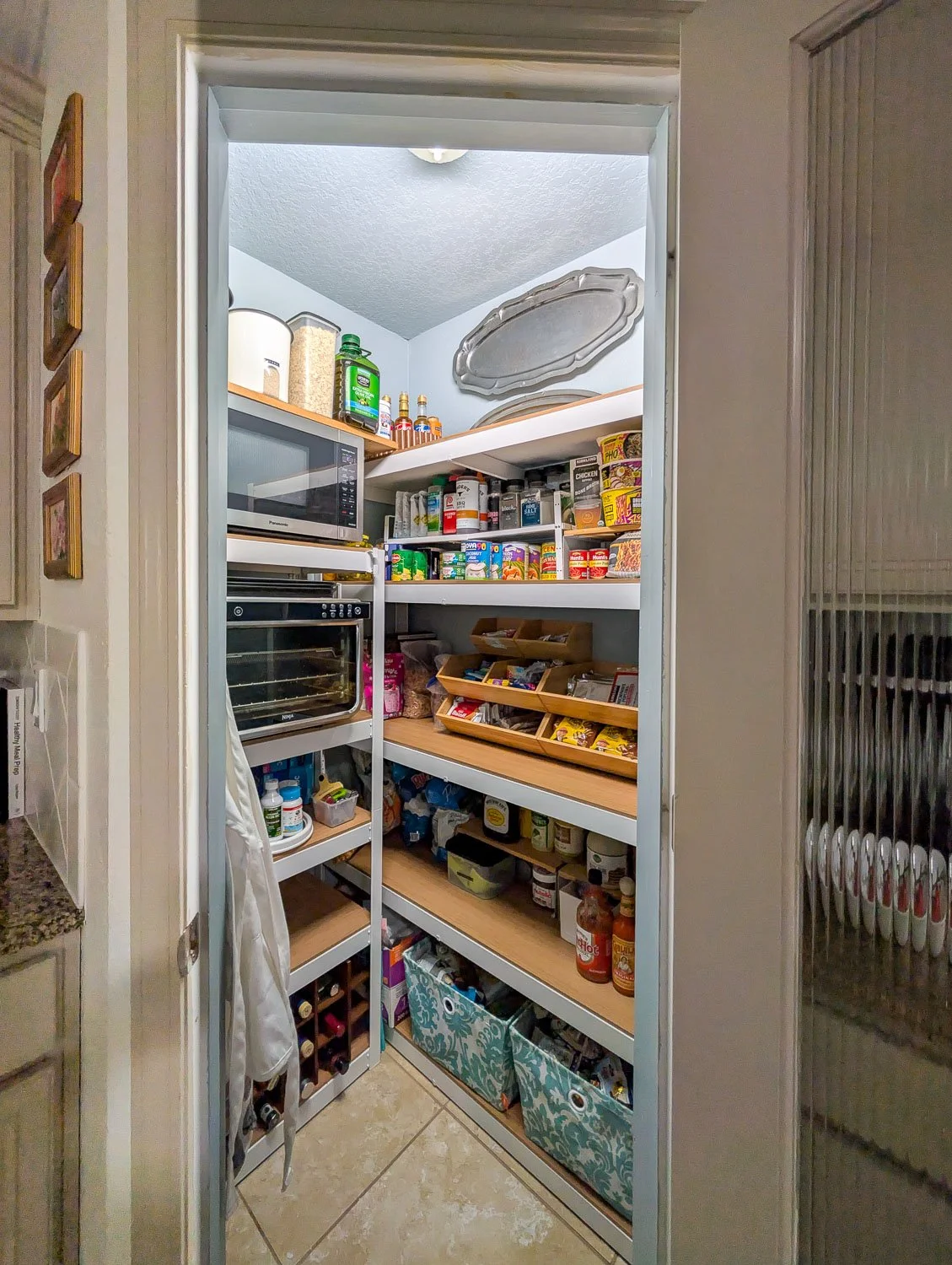 A remodeled walk-in pantry painted light blue, featuring sturdy white metal shelving with wood liners, organized food items, and decorative silver platters on the wall.
