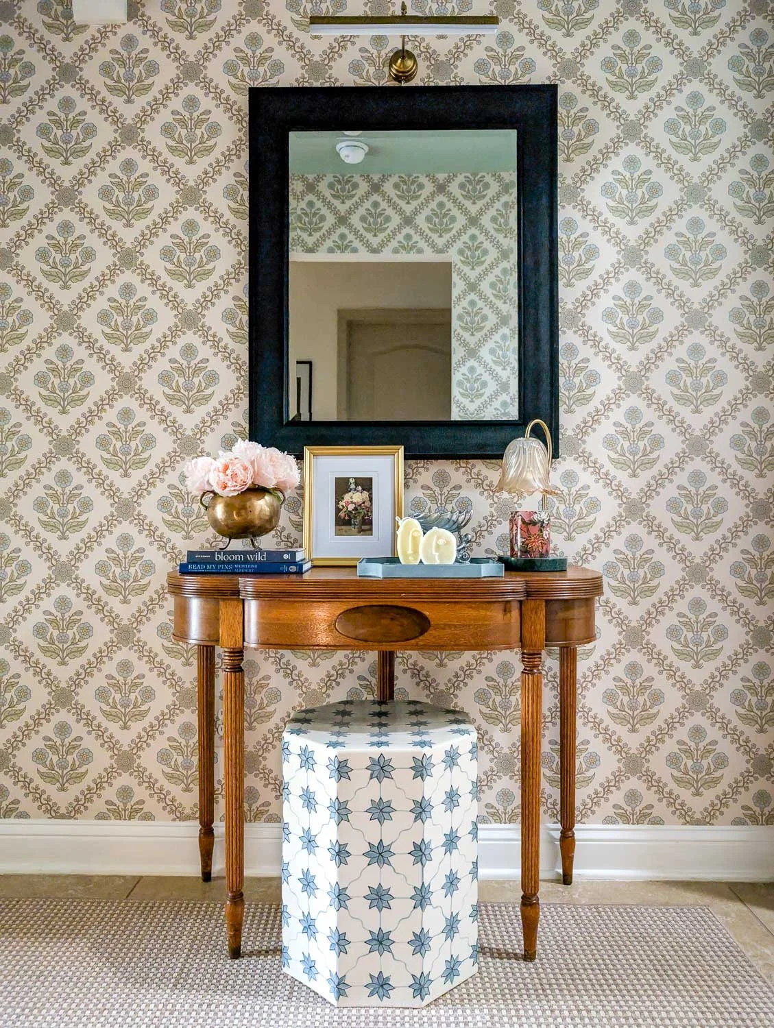 A traditional wooden console table decorated with pink flowers, framed art, and small accents sits against patterned floral wallpaper beneath a black-framed mirror, with a blue-and-white patterned ceramic stool tucked underneath.