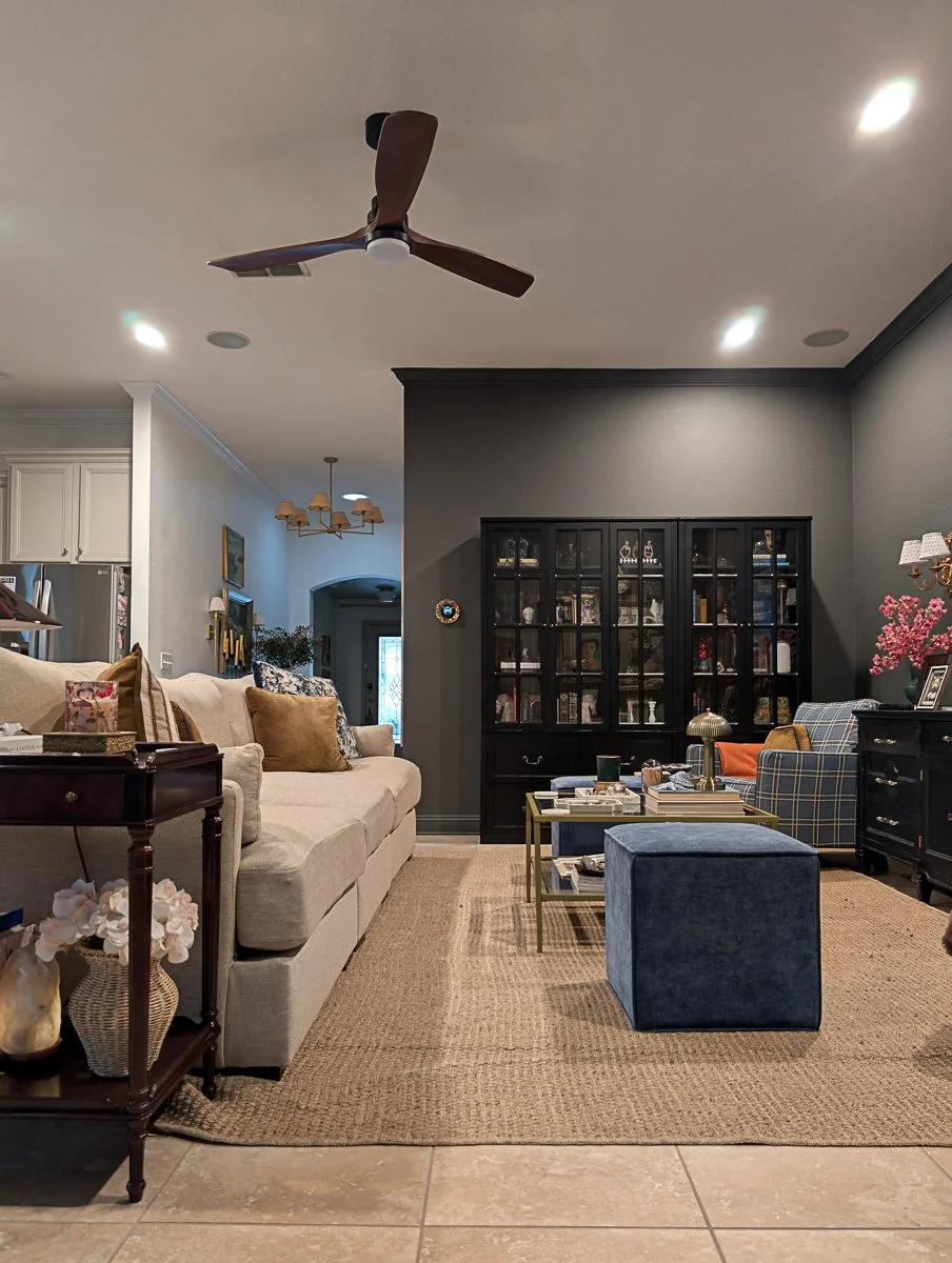 A bright, open-concept living space showing a transition from a neutral-toned hallway to a moody sitting area with dark walls, a jute rug, and layered lighting from recessed ceiling lights and a central fan.