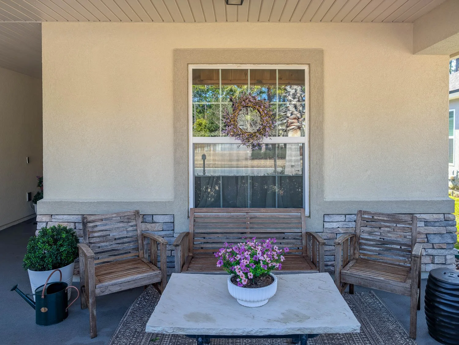A cozy front porch sitting area features weathered wooden furniture, a stone-topped coffee table with purple flowers, and a decorative wreath hanging on the window.