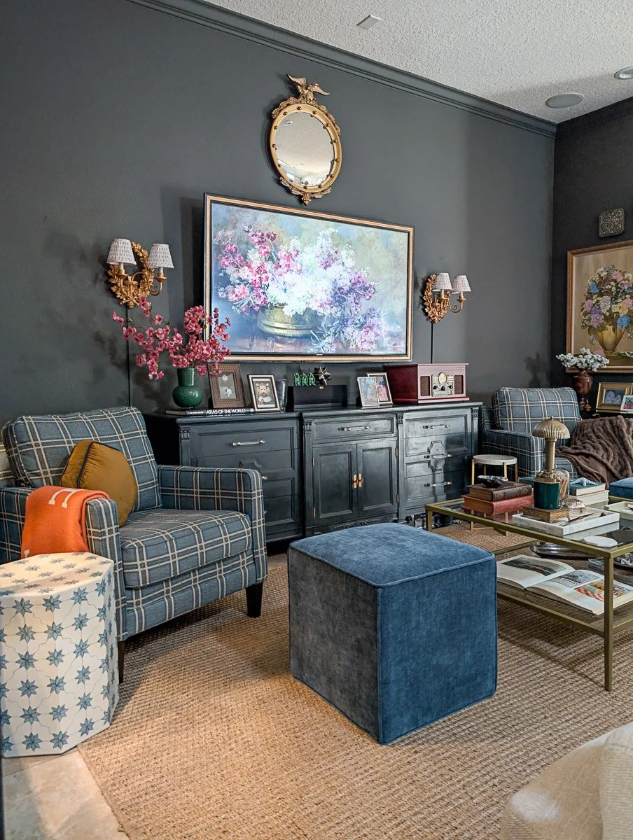 A moody, traditional living room features green walls, blue plaid armchairs, and a large floral screensaver with TV mounted above a black vintage sideboard.