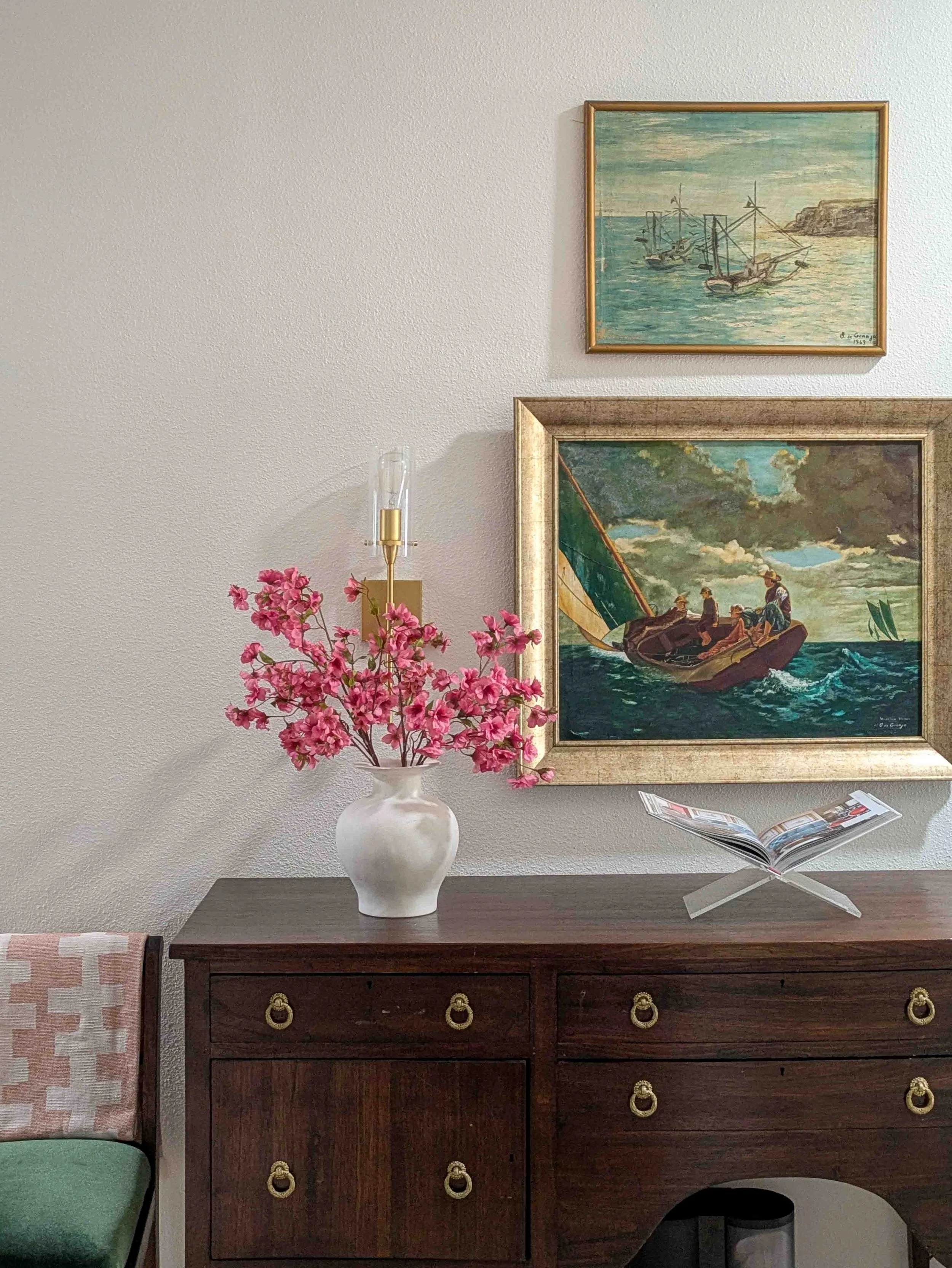 A dark wood vintage dresser styled with a white vase of vibrant pink cherry blossom branches, an acrylic book stand, and a gallery wall of coastal oil paintings.
