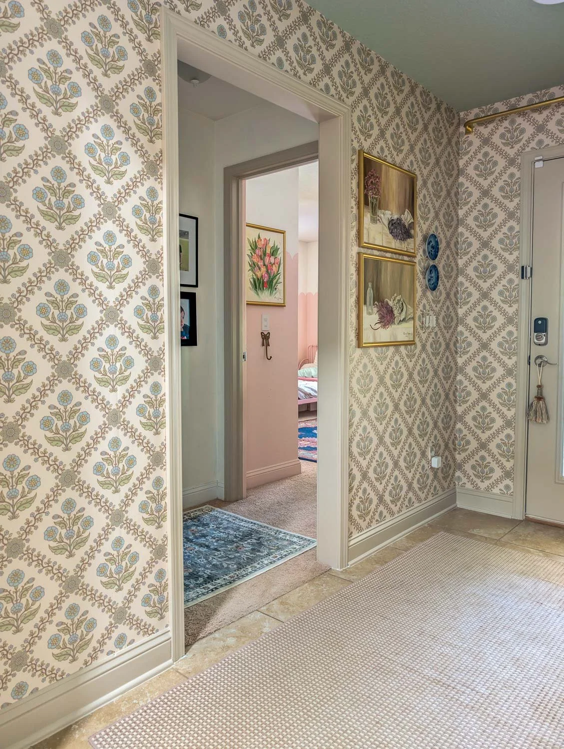 An angled view of an interior hallway showing the transition from a wallpapered entryway to a neutral-painted corridor with framed artwork.