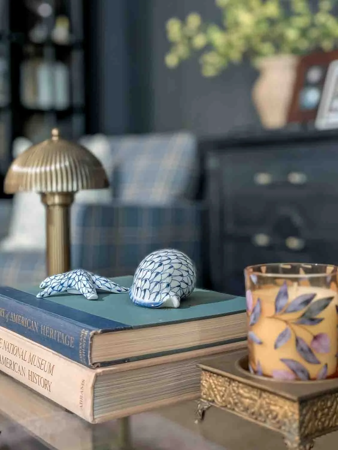 A close-up, shallow-depth-of-field shot of a glass coffee table. A stack of vintage hardcover books sits in focus, topped with blue and white patterned porcelain figurines of a starfish and a shell.