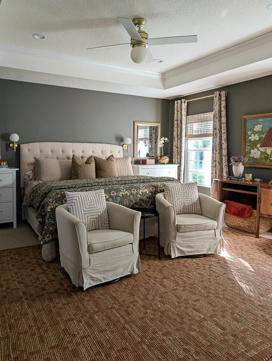 A sophisticated primary bedroom features dark olive-green walls, a tufted cream headboard with a floral quilt, and two matching beige armchairs positioned at the foot of the bed on a large woven jute rug.