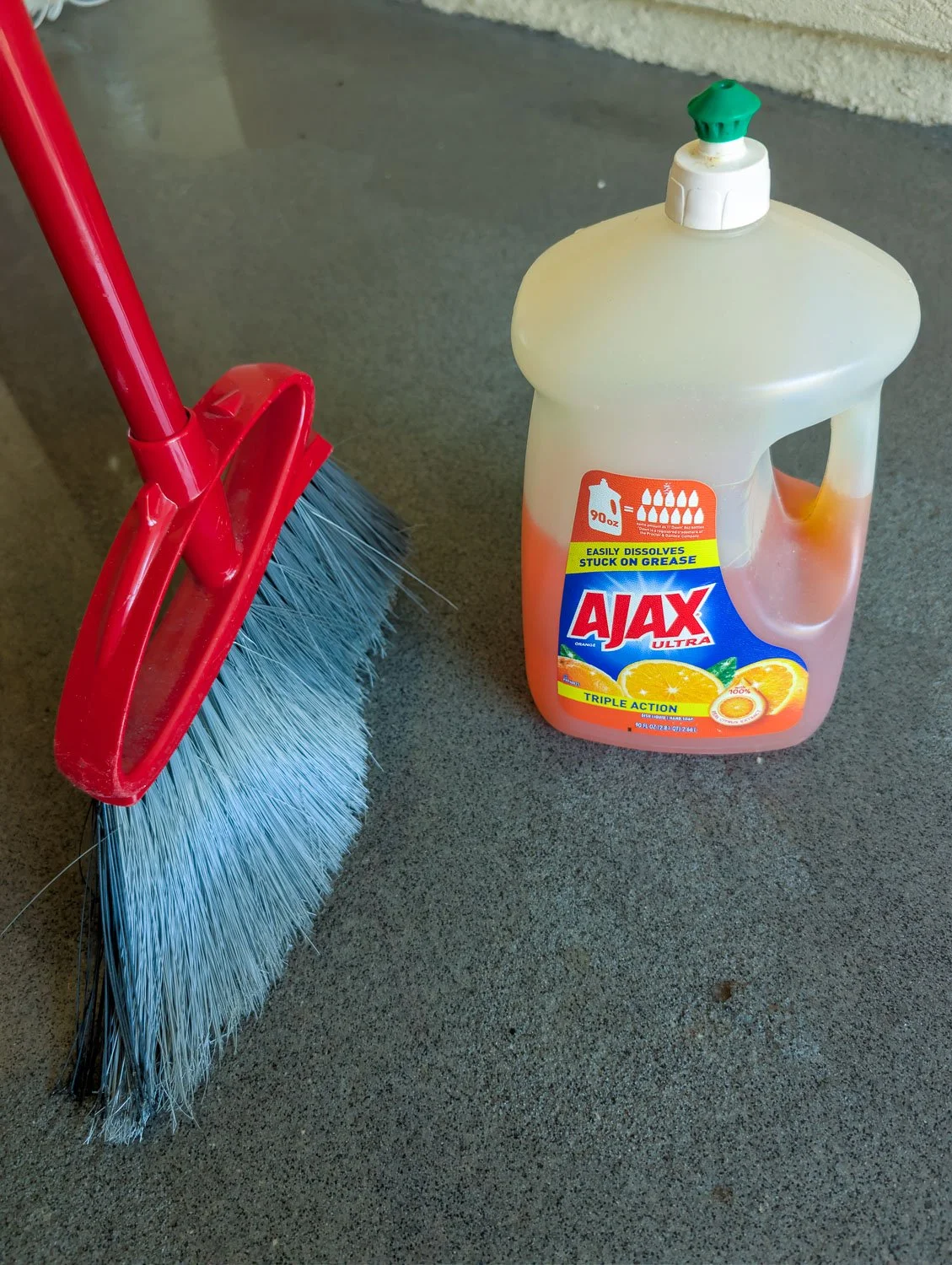 A red broom with gray bristles stands next to a large bottle of orange Ajax Ultra dish soap on a gray concrete surface.