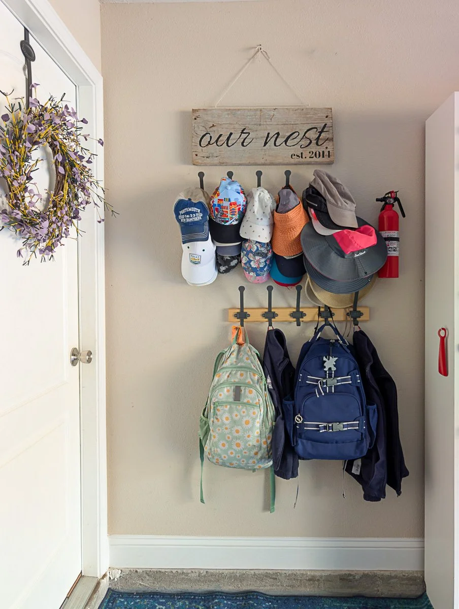 Two wooden racks are mounted on a beige wall, heavily loaded with various colorful hats, backpacks, and jackets next to a white door.