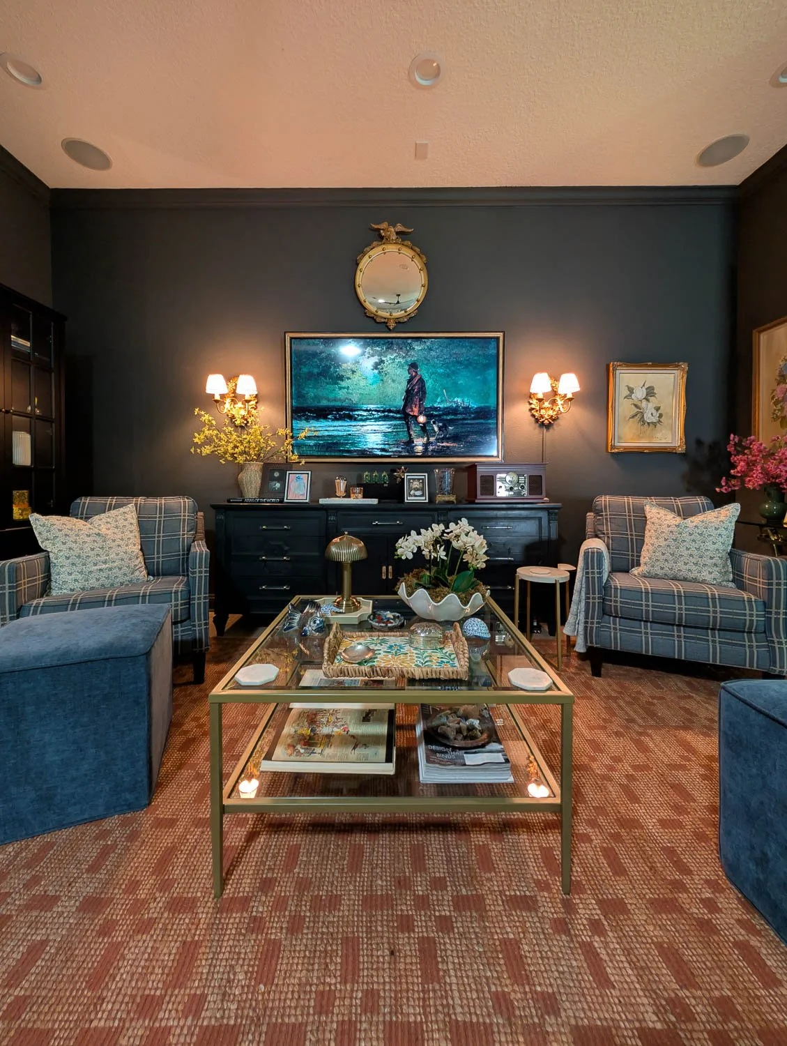 A symmetrical living room arrangement showing a black media console under a large glowing beach painting, illuminated on either side by warm, ornate brass wall sconces against dark charcoal walls.