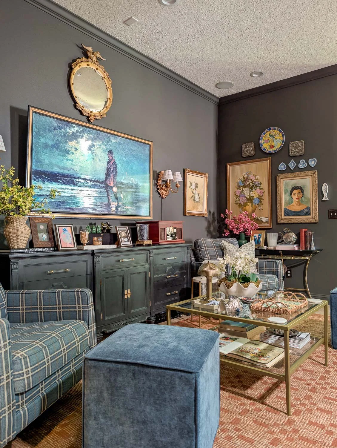 A beautifully styled modern heritage living room featuring dark gray walls, blue plaid armchairs, a terracotta area rug, and an elegant console table setup underneath an illuminated coastal landscape painting.