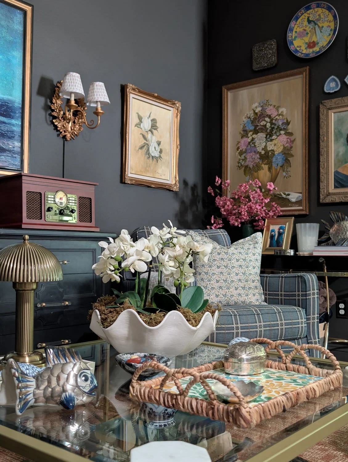 Close-up of a coffee table vignette with a scalloped white shell bowl of orchids, a fluted brass dome table lamp, and decorative metallic fish, with an ornate brass wall sconce and dark gray gallery wall in the background.