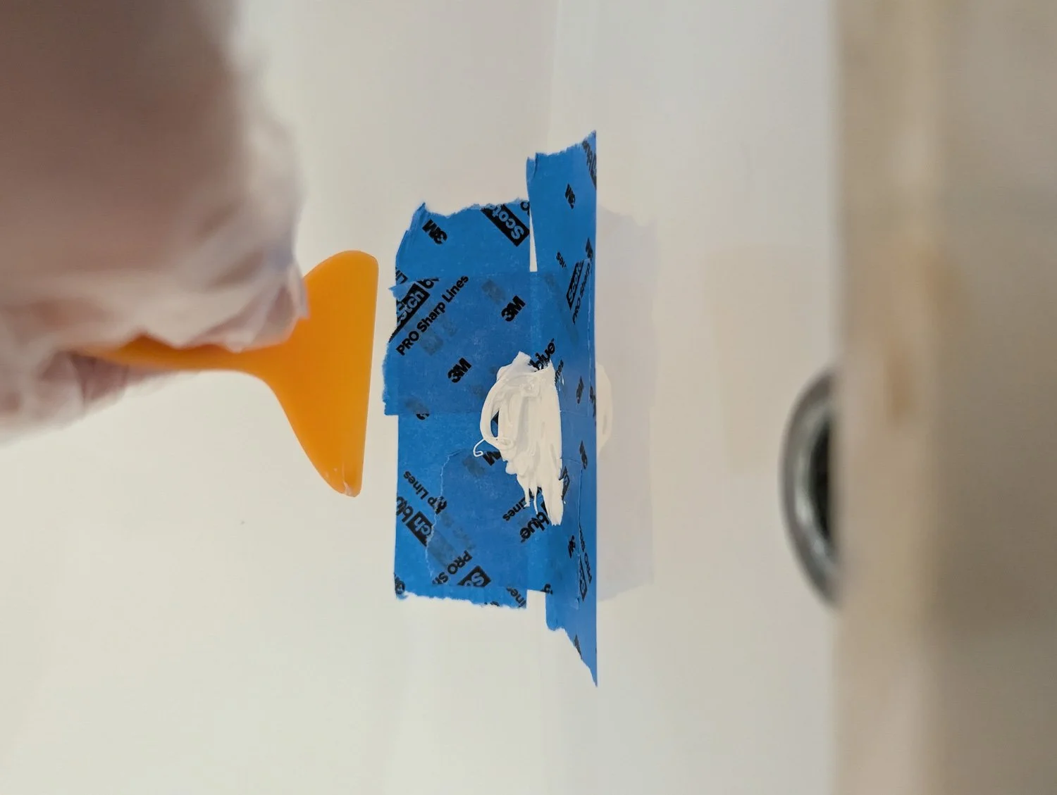 A close-up of a small chip in a white porcelain surface, isolated and surrounded by strips of blue painter's tape to protect the surrounding area.