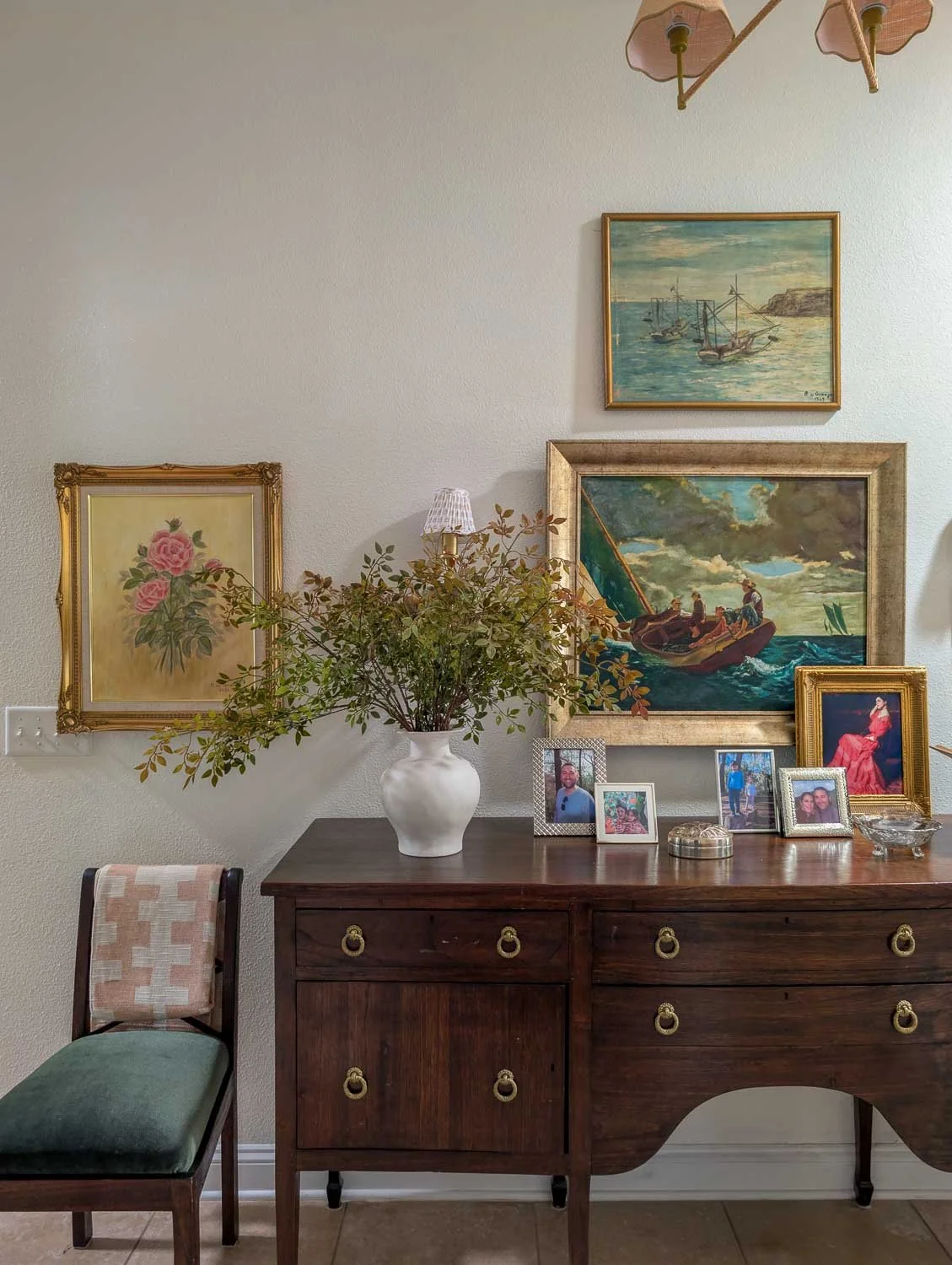 A dark wood antique sideboard decorated with gold-framed maritime and floral oil paintings, family photos, and a white vase with leafy branches against a neutral wall.