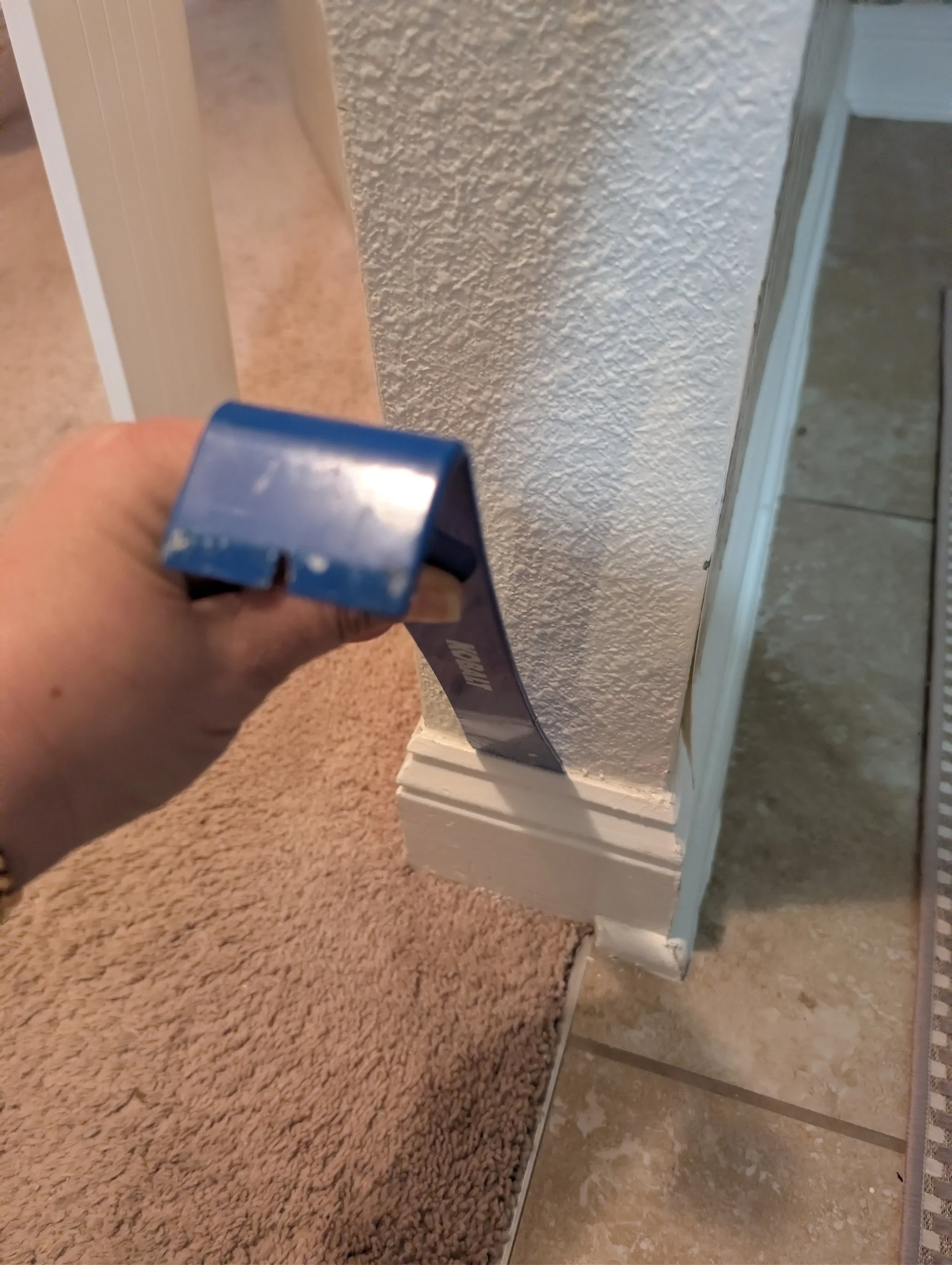 A person uses a blue pry bar to carefully remove a section of white baseboard from a textured wall.