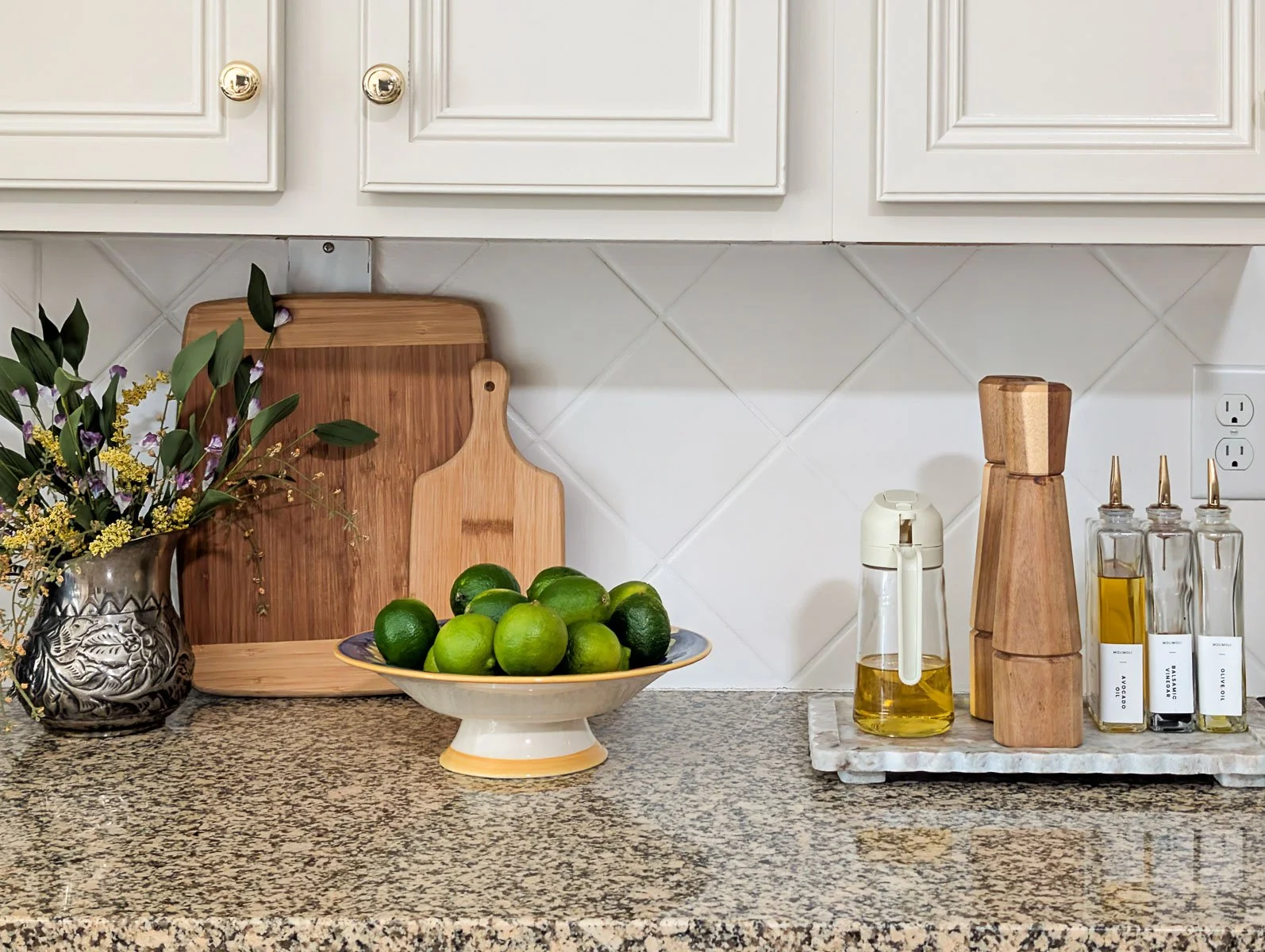A close-up of a kitchen countertop shows a bowl of fresh limes, wooden cutting boards, and glass oil carafes arranged against a white diamond-tile backsplash.