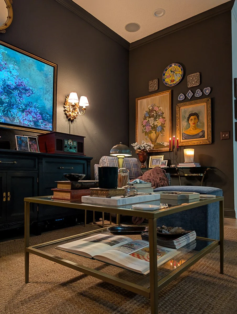 A cozy, maximalist living room featuring dark charcoal walls adorned with a gallery of vintage paintings and a gold sconce, centered around a brass coffee table with books and a blue velvet ottoman.