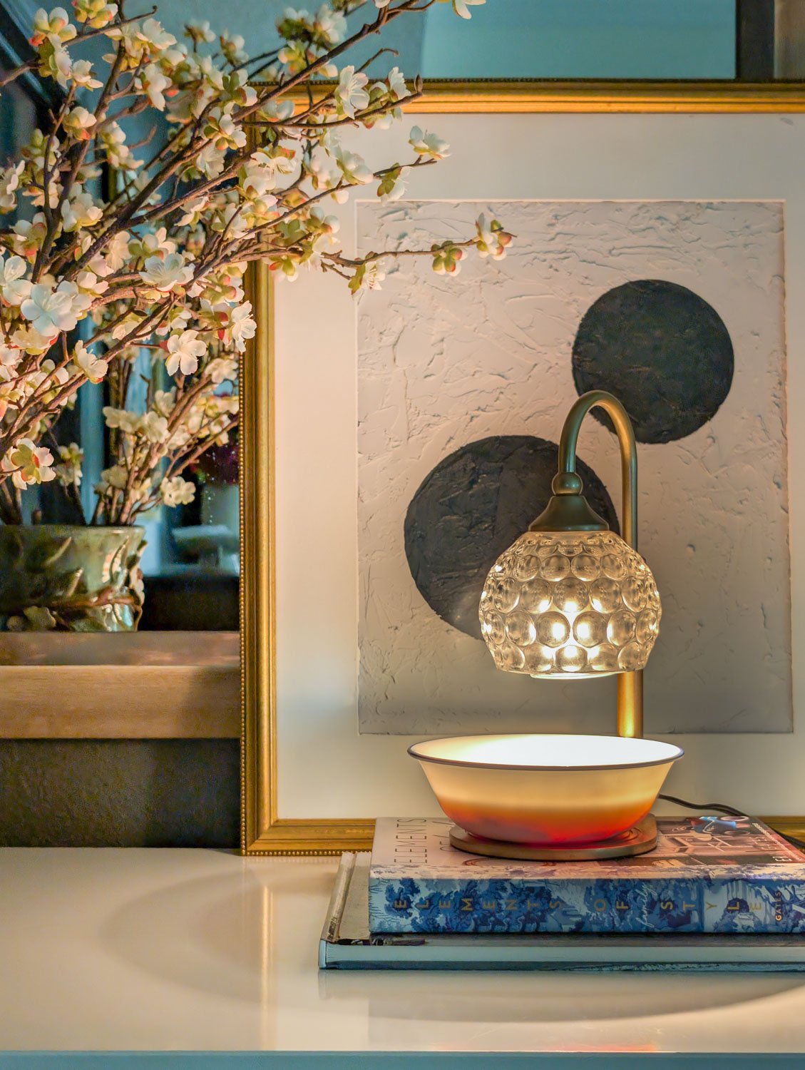 A gold-finished candle warmer lamp illuminating a white bowl of natural simmer pot ingredients on a bedroom dresser.