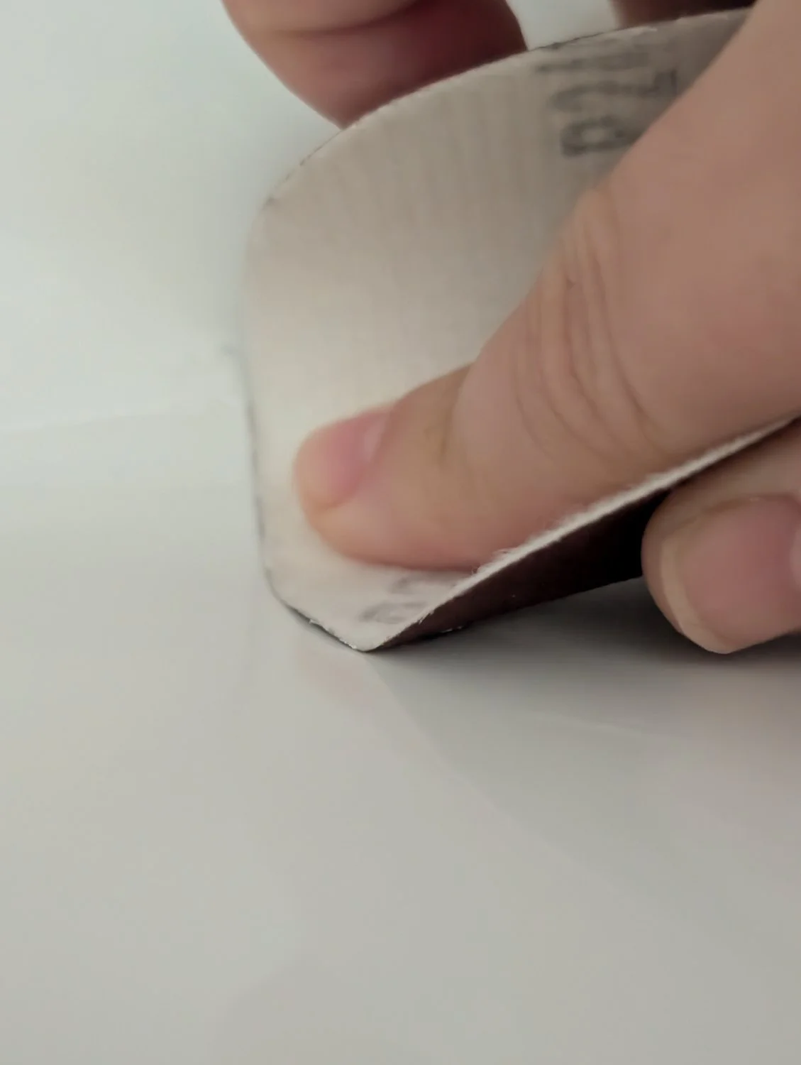 A hand using a piece of fine-grit sandpaper to gently smooth down a cured white epoxy patch on a porcelain sink surface.